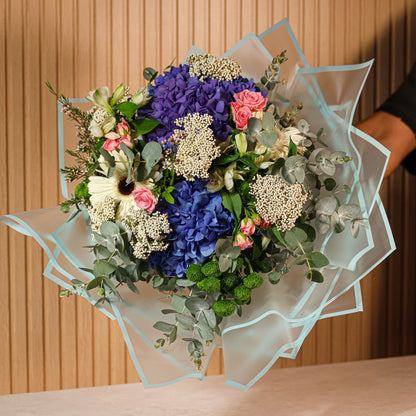 Ramo de flores preservadas Aqua Belle con hortensias azules, gerberas blancas, rosas spray rosadas y eucalipto verde